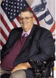 A man wearing glasses, a dark blazer, maroon sweater, and pink tie sits in a wheelchair, smiling. Behind him are the American flag and another flag with partial text and a graphic.